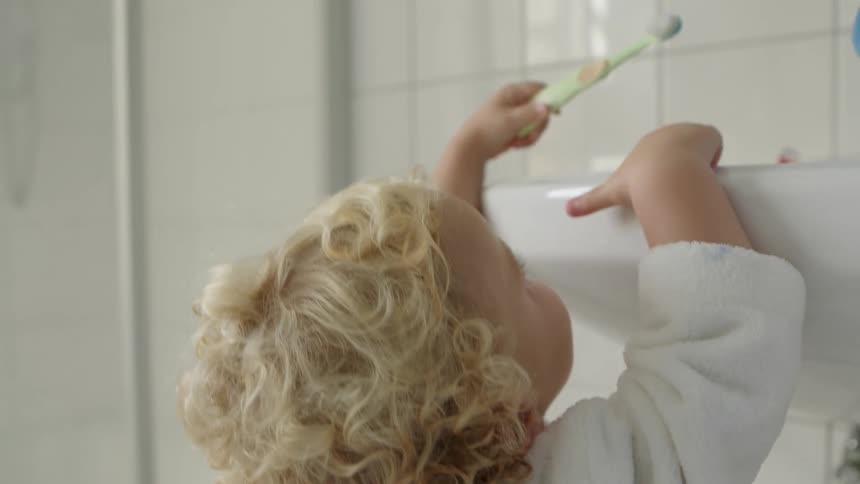 A blonde child is in the bathroom reaching for the sink holding a toothbrush