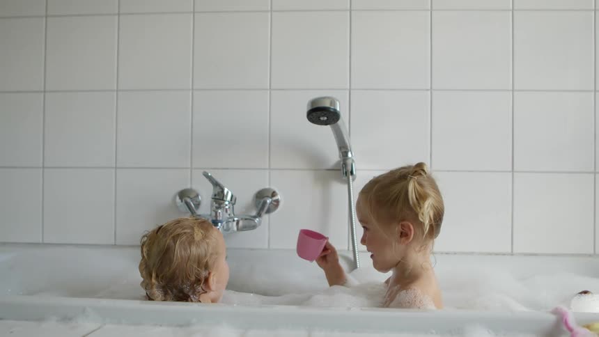 Two blonde girls are in a bubble bath in a white tub with white tiles