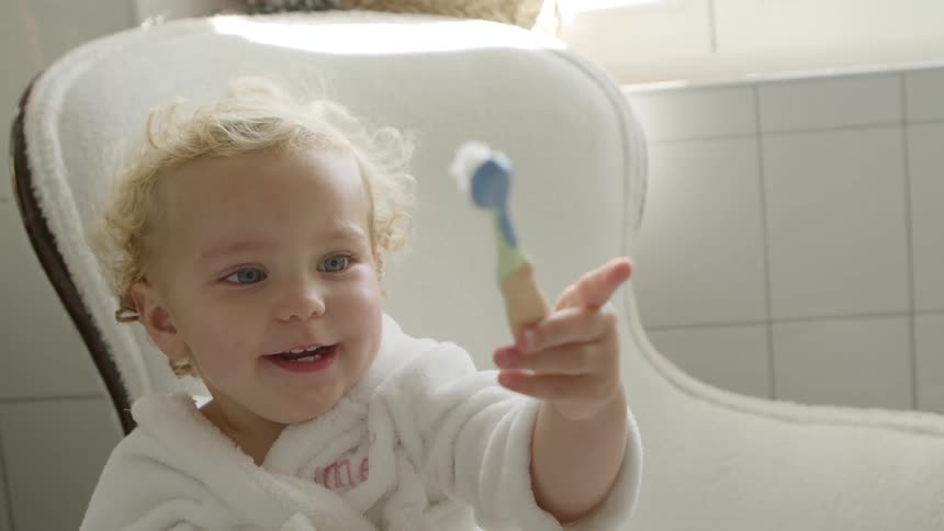 Blonde toddler girl wearing a white bathrobe sits in a white chair and brushes her teeth in the bathroom
