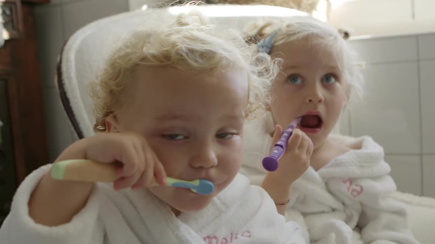 Two baby girls sitting in the bathroom wearing bathrobes brushing their teeth