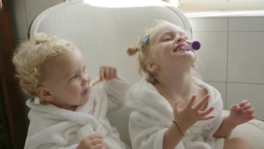 Two toddler girls sit in the bathroom wearing robes and have toothbrushes in their mouth