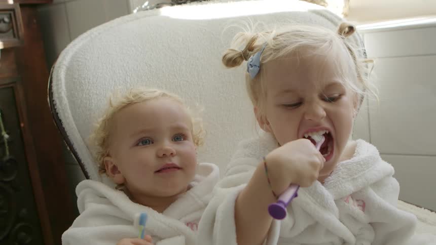 Two siblings sit on a chair together in the bathroom and brush their teeth