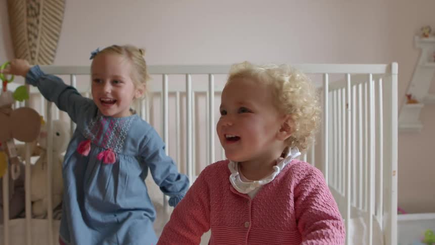Two blonde girls in a baby room next to a white crib at home