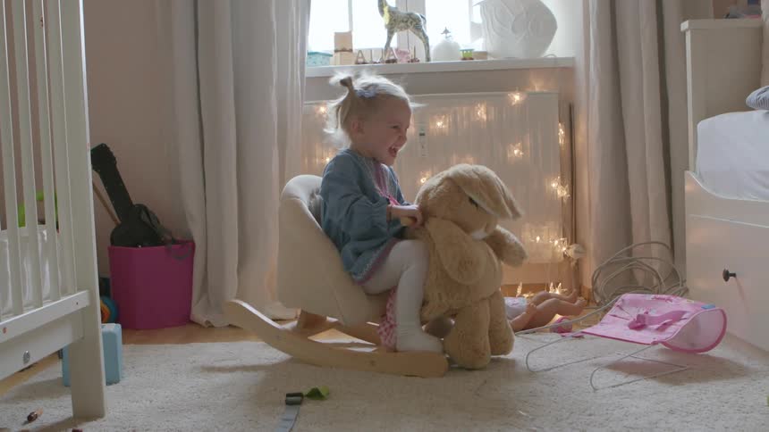 A little girl laughs and smiles as she rides her rocking bunny in her bedroom