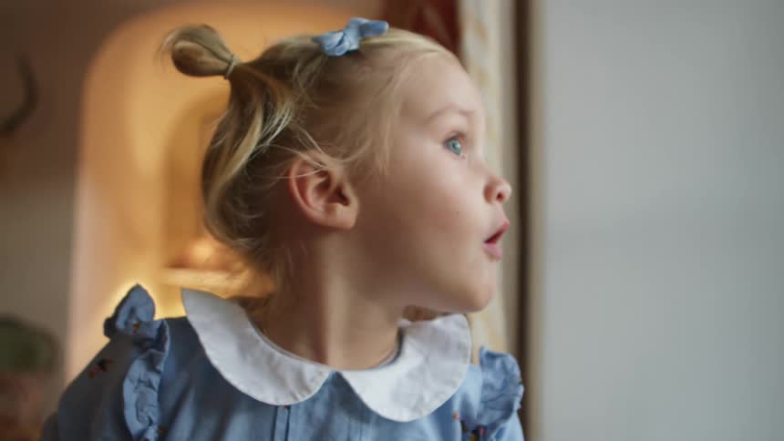 Blonde toddler girl with a light blue dress looks out the windows at home