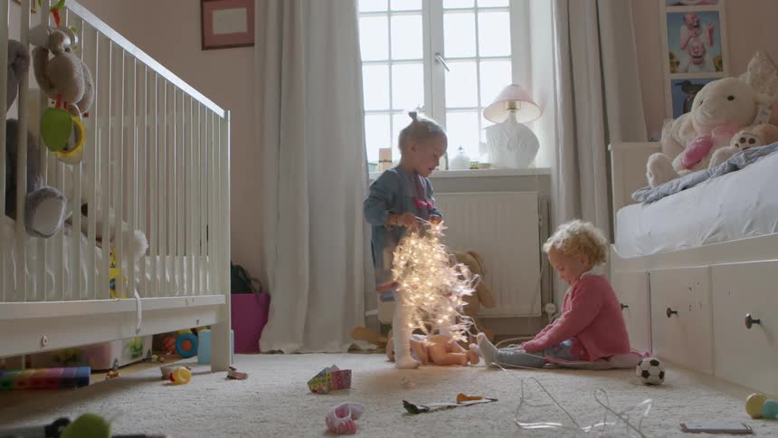 A young girl sitting on the carpet of a childs bedroom rises to take a string of fairy lights away from a younger girl sitting across from her