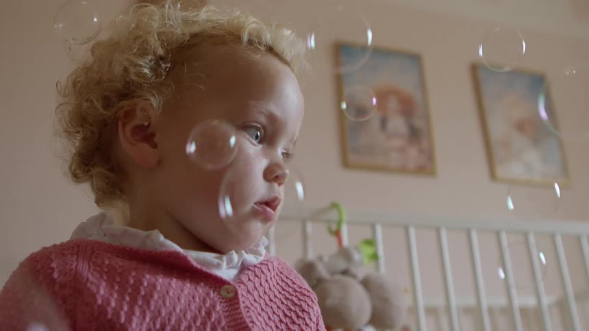 A blonde toddler stands in a room with a white crib and bubbles floating in the air