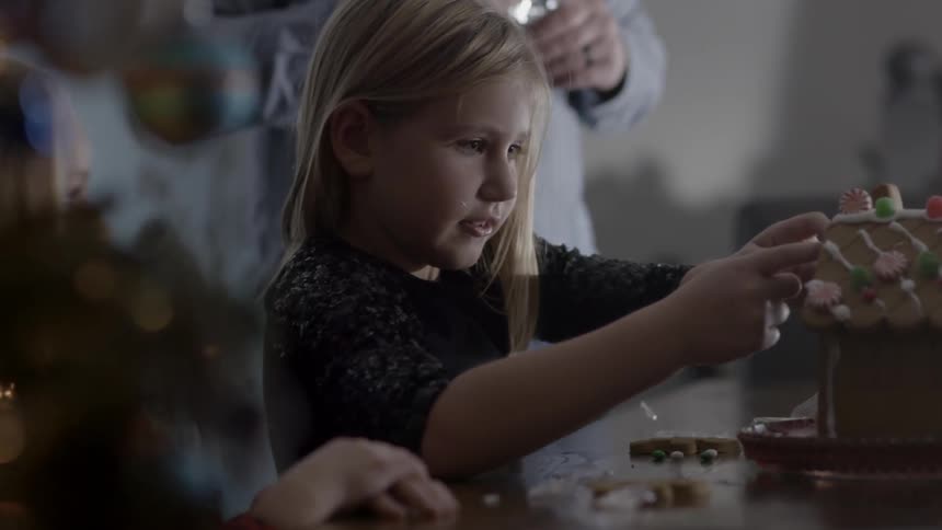 A young girl decorates a gingerbread house while a parent supervises