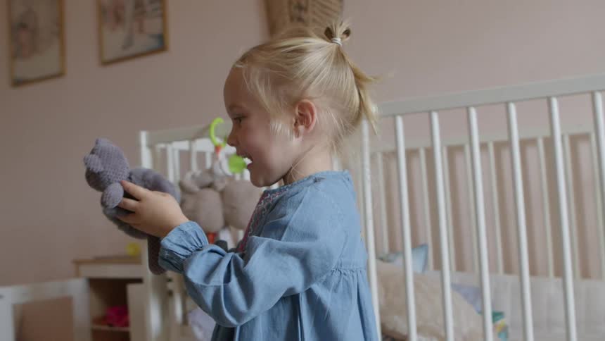 A young girl plays with a small stuffed animal while walking next to a crib in a bedroom
