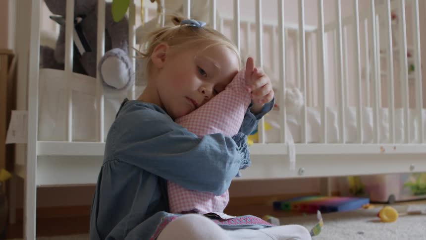 A young girl sitting on the floor in front of a crib in a bedroom hugs a cushion to her face