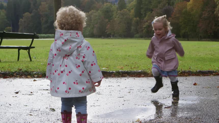 One little girl runs and jumps in a puddle splashing another girl who is standing near by in a park on a cloudy day in the fall with a bench in the grassy area in front of the trees in the background
