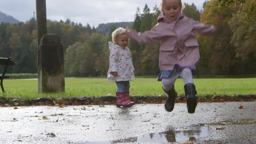 A girl with rain boots jumping on a puddle with trees the sky and clouds in the background