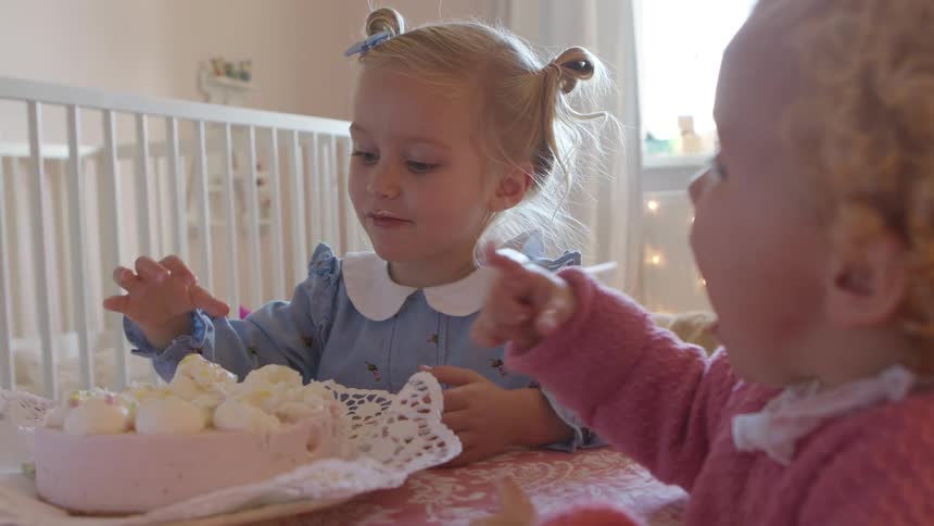 Two young girls sit at a table in a bedroom eating ice cream with their hands and licking their fingers