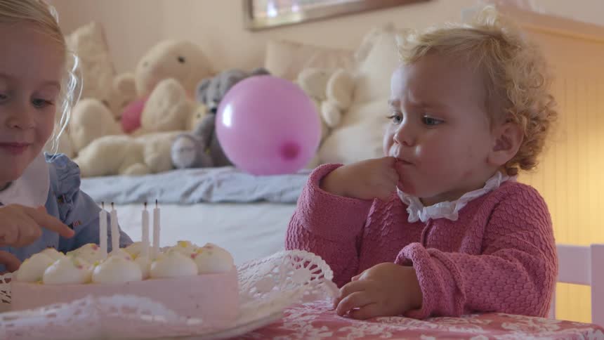 A pair of young girls sitting at a childrens table in a childs bedroom stick their fingers into the icing of a cake before sticking their fingers in their mouth