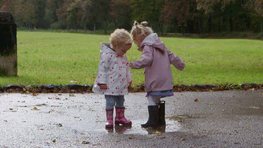 Two young youngs jump into a driveway puddle wearing rain coats and rain boots