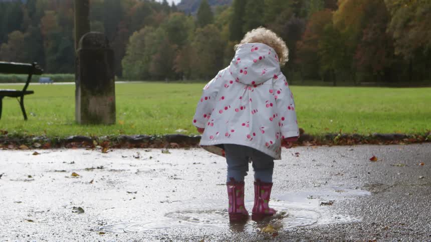 A young girl in a raincoat and rain boots plays in a puddle on a walkway in a park during the day