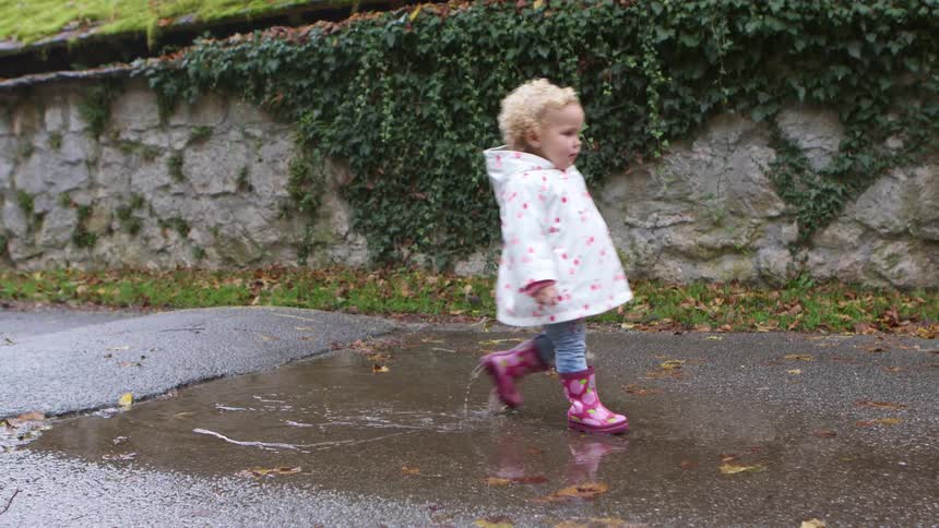Blonde toddler girl with a white coat and pink boots walks through a puddle in the street