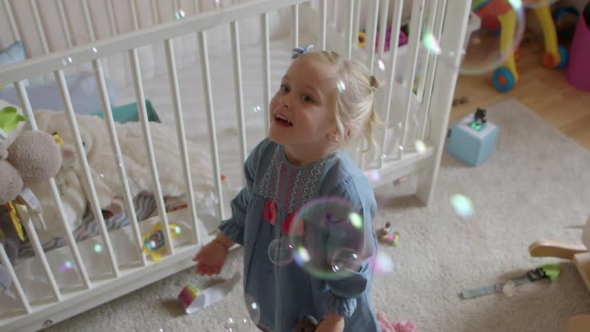 Little girl standing in a babys room playing with bubbles being blown at her