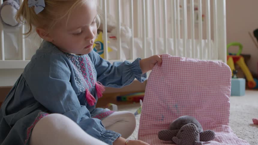 Little girl in a blue dress playing with a teddy bear in her room next to a crib