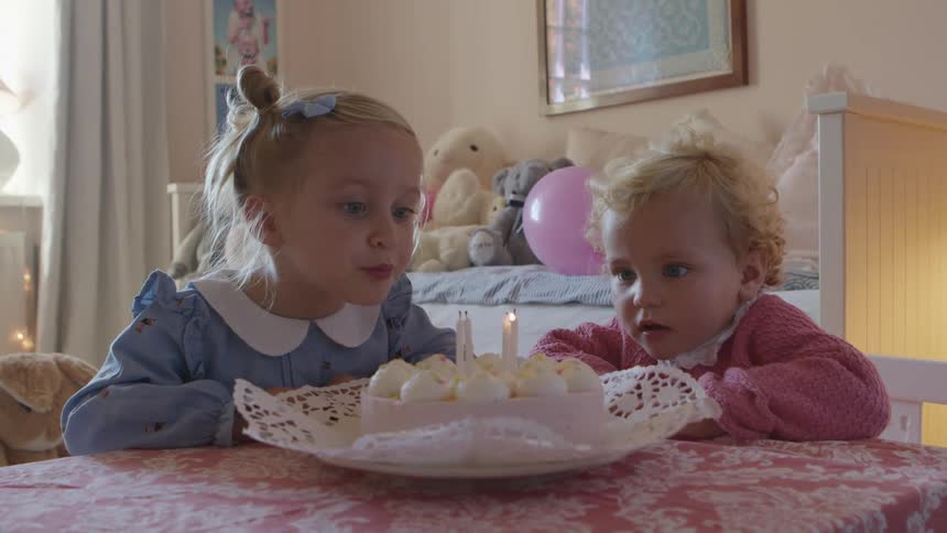 Two girls sitting at a table in a room as one girl blows out the candles on a birthday cake