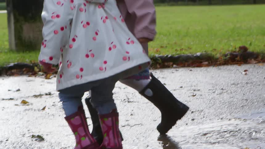 A young girl in raincoat runs and jumps next to her sister in a puddle on a walkway in a park