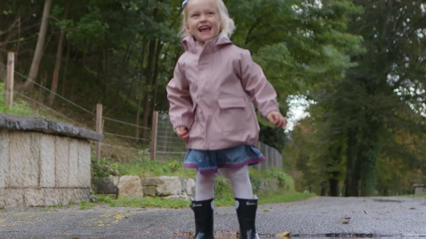 A young girl wearing a raincoat and rain boots jumps into a puddle in a gravel driveway