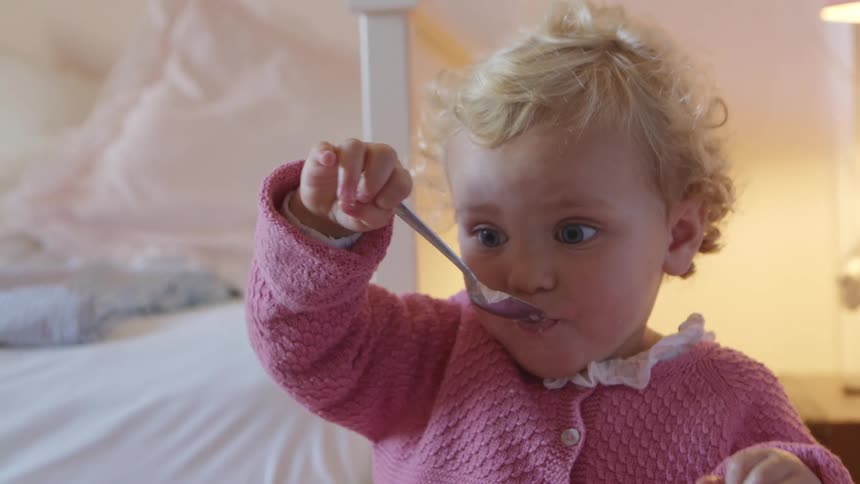A toddler with blonde hair eagerly feeds herself food using a spoon