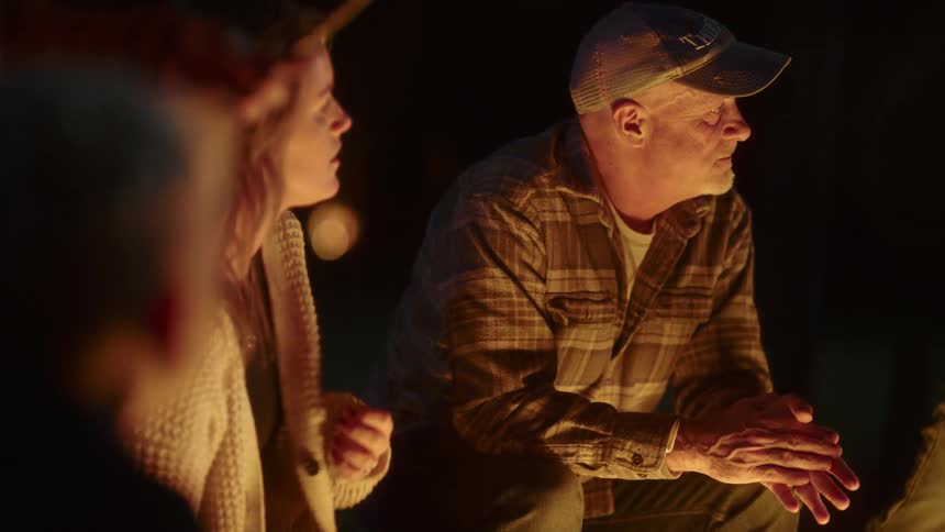 A man sits in front of a fire outdoors surrounded by other people at night