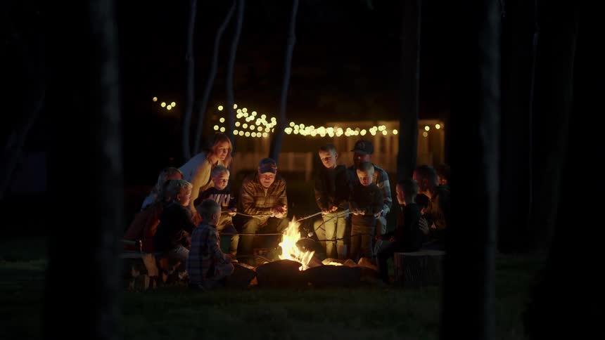 A group of men women boys and girls toast marshmallows on a campfire at night