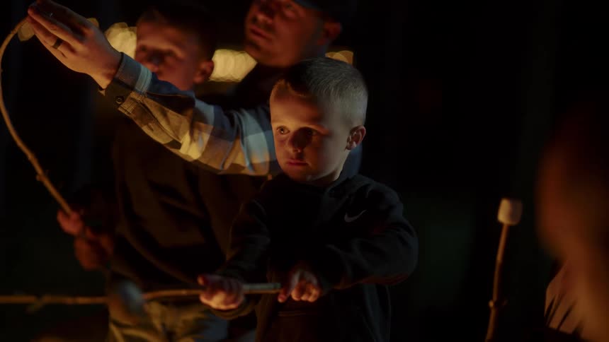 A father and his sons outside in front of a campfire roasting marshmallows on a stick at night time