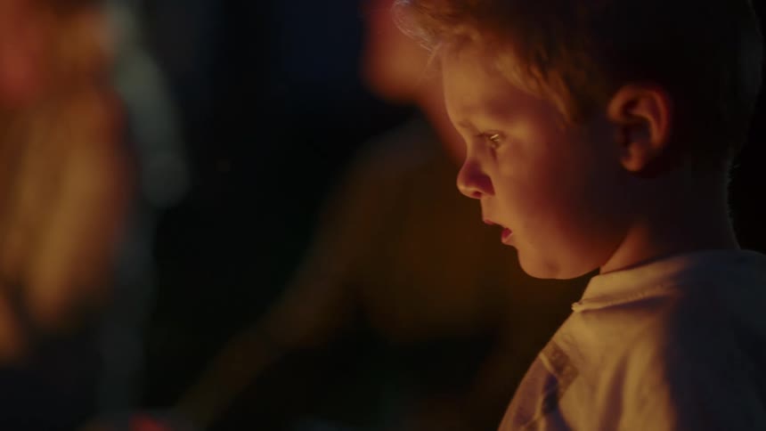 Blonde boy looking at a campfire with family at night