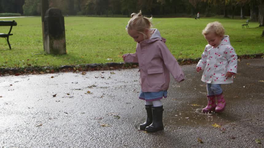 A young girl in a raincoat jumps in a puddle in a park and is joined by her toddler sister