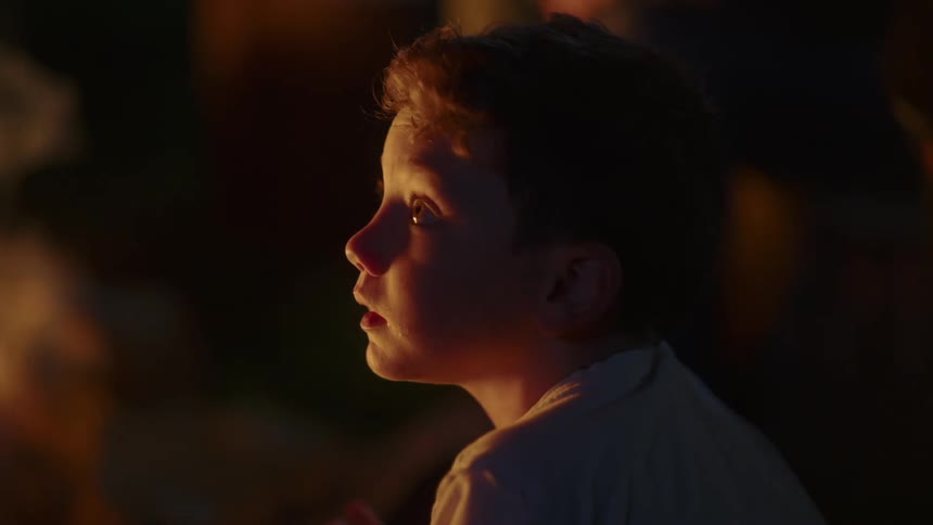 Boy sitting down in front of a flickering light looks up and around him at nighttime