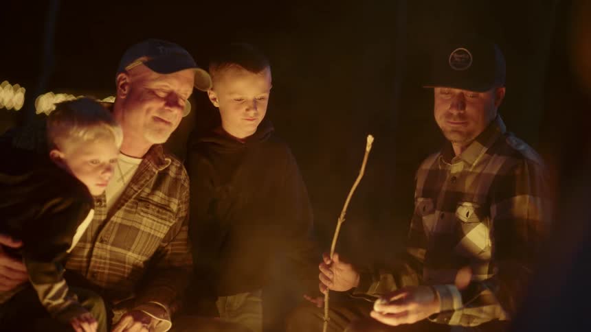 A man holds his grandson as he watches his son help another boy roast a marshmallow over a campfire