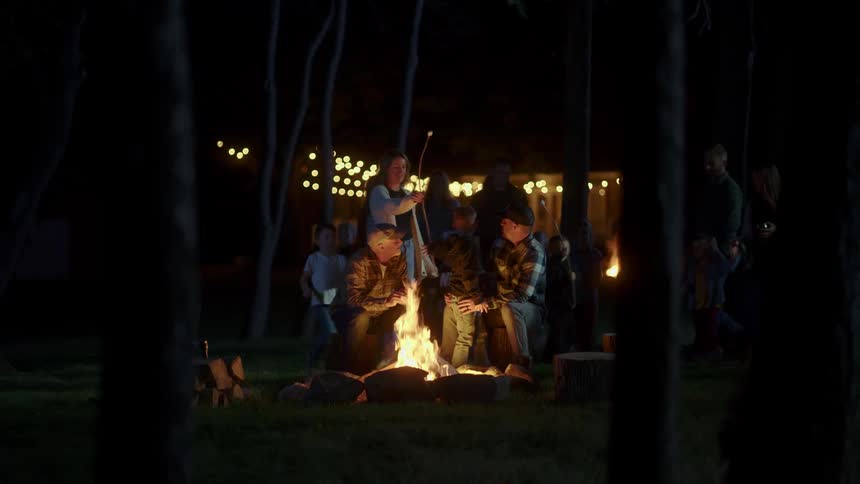 A woman brings a marshmallow on a stick over to her husband and son who are roasting marshmallows over a bonfire