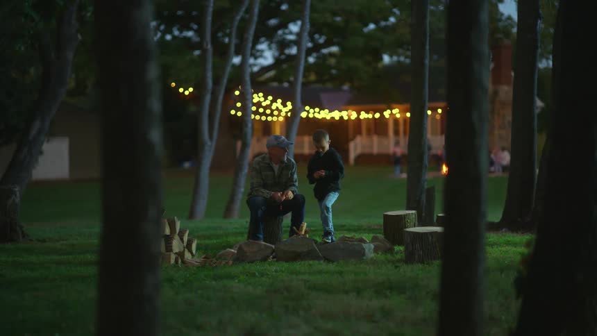 A man sitting on a tree stump in front of an unlit fire circle talks to a boy standing next to him