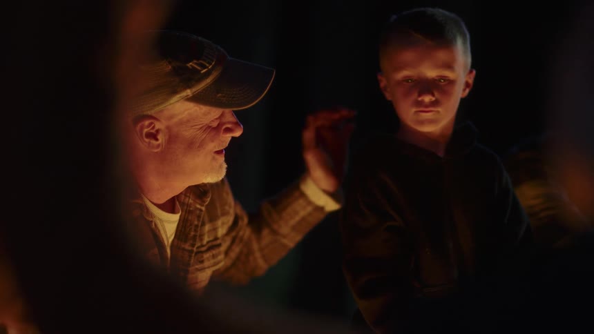 Two men and a boy with a stick in his hand around a campfire smiling