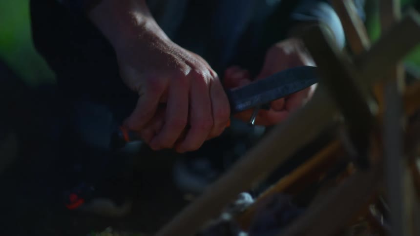 A man kneeling before a stack of wood scrapes a knife against a flint to create sparks in an attempt to start a campfire