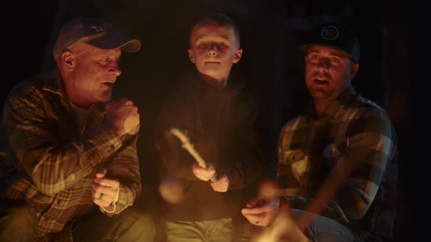 Two men and a boy by a campfire roasting a marshmallow on a stick with trees in the background