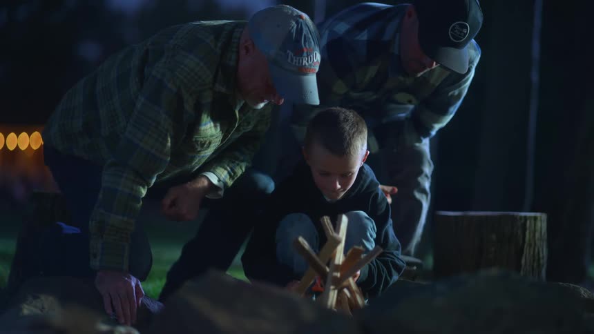 Two men kneel beside a boy trying to start a campfire