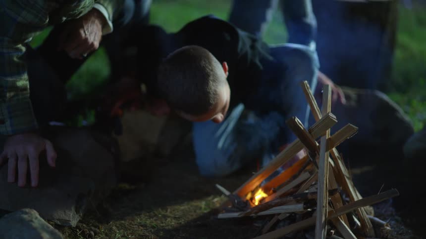 Boy sitting in front of a small not fully lit campfire blowing the flames out at night time