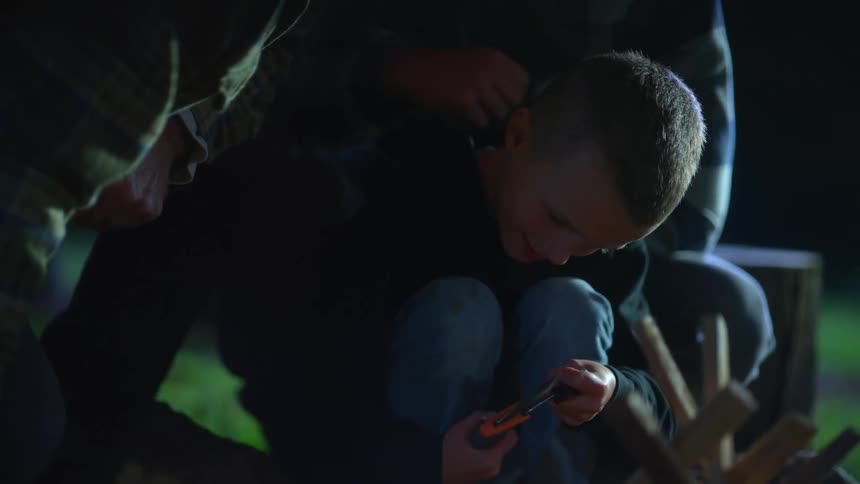 Young boy sitting in front of his father outside at nighttime with a carving knife scraping it against an object in his hand