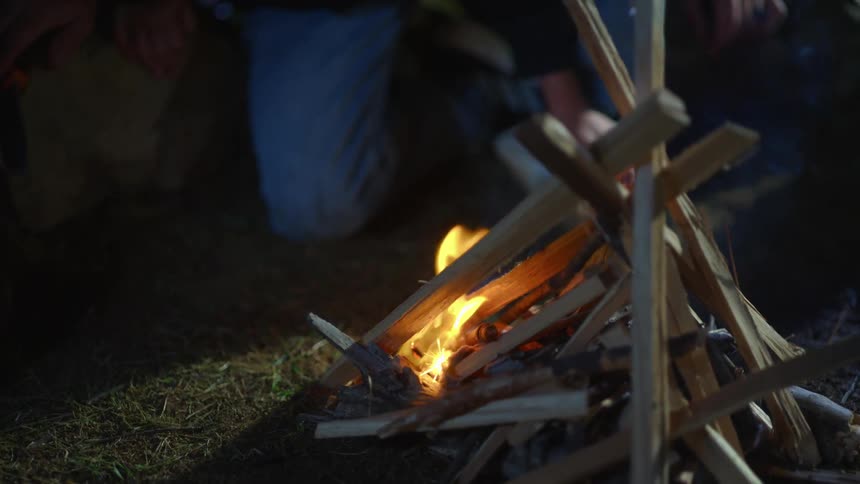 Boy knelt down in front of a pile of sticks trying to keep a campfire going