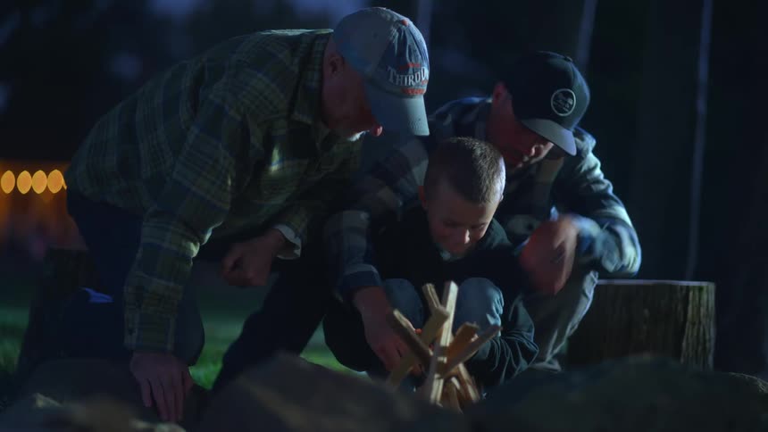 Two men kneel next to a boy attempting to build a campfire at night
