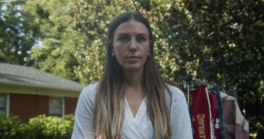 Woman with brown hair and a white shirt faces the camera with eye contact in the garden