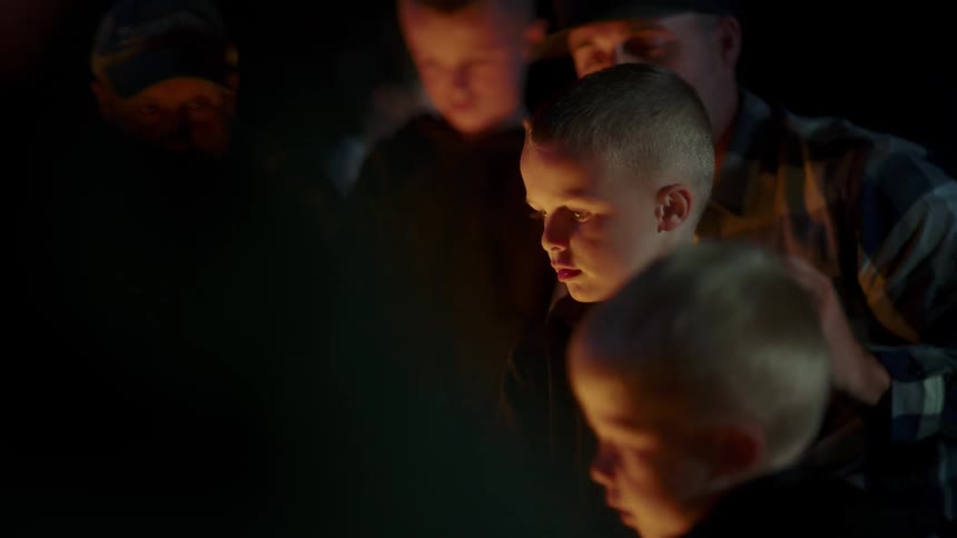 A son sits in his dads lap as they sit around a fire outside with other children and an adult at night