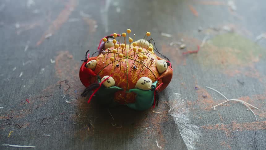 Several sewing pins are stuck in a pincushion with small people as decorations along its sides
