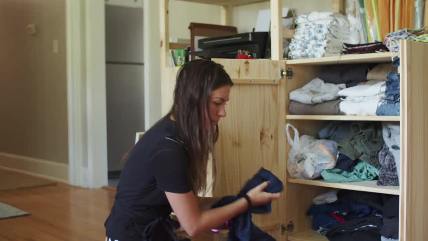A woman sits on a hardwood floor with a cabinet open sorting through clothes during the day