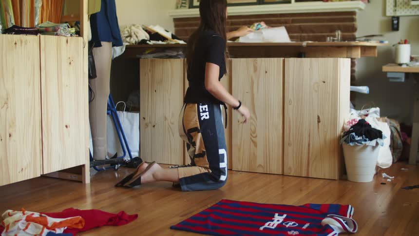 A young woman kneeling in a room spreads a soccer jersey on the floor before retrieving paper from a nearby shelf
