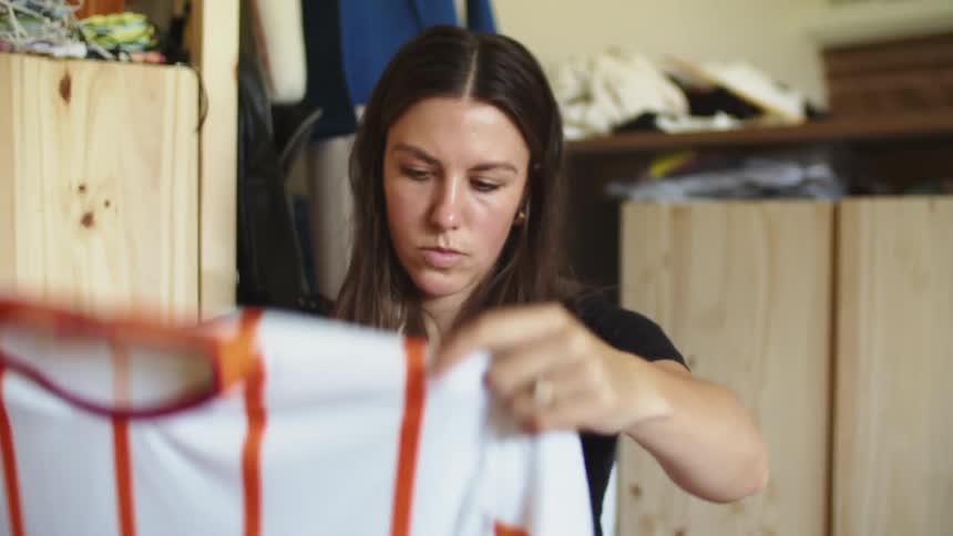Woman sitting down in a room in front of a wooden stand folding an orange and white jersey shirt